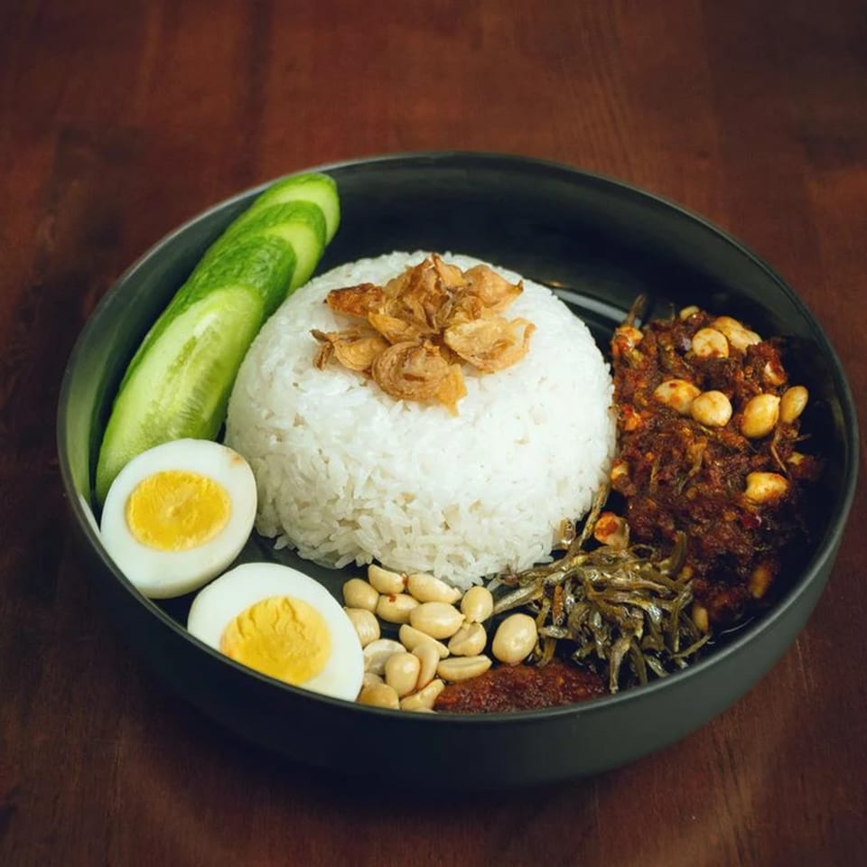 Nasi Lemak with rice, fried anchovies, peanuts, sambal, cucumber, and hard-boiled eggs in a black bowl.