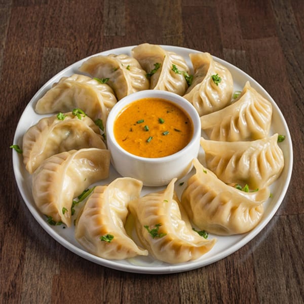 A white plate of steamed momos with a small bowl of orange dipping sauce in the center.