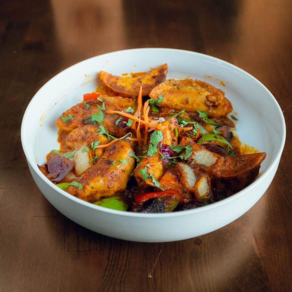 Spicy potato wedges with bell peppers, onions, and cilantro in a white bowl on a wooden table.