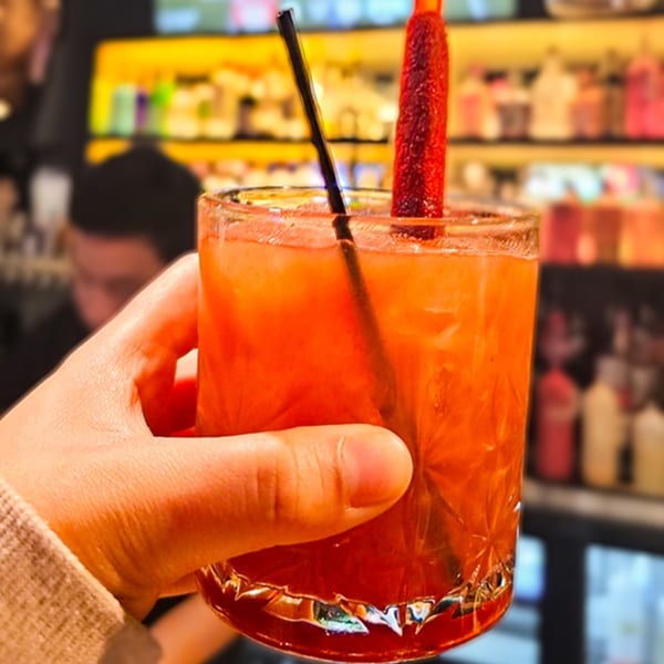 A hand holds a vibrant orange cocktail with a black straw and a red candy stick.