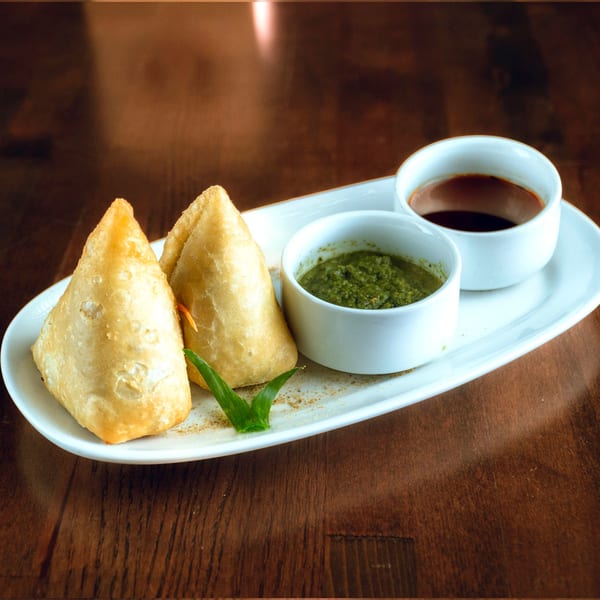 Two golden-brown samosas with green and brown chutneys on a white platter.