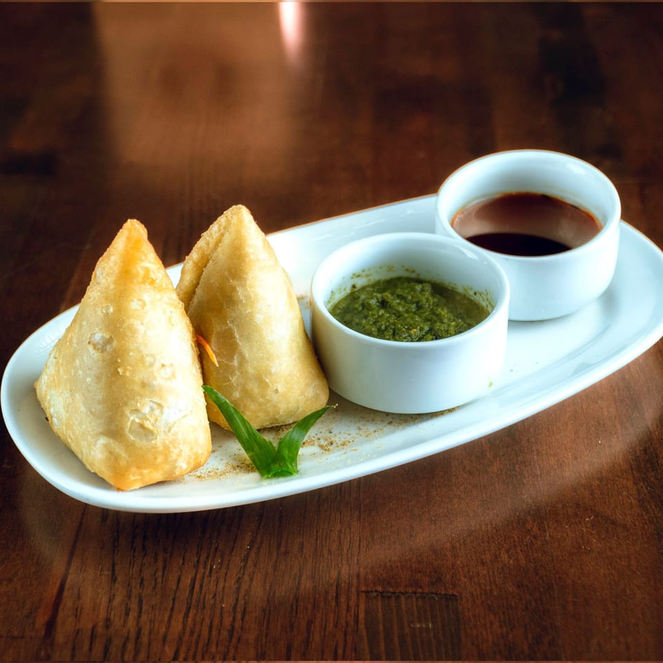 Two golden-brown samosas with green and brown chutneys on a white platter.