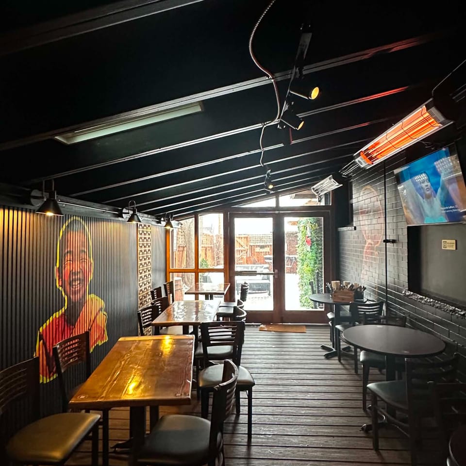 Dark restaurant patio with wooden tables, a smiling child mural, TV, heaters, and glass doors.