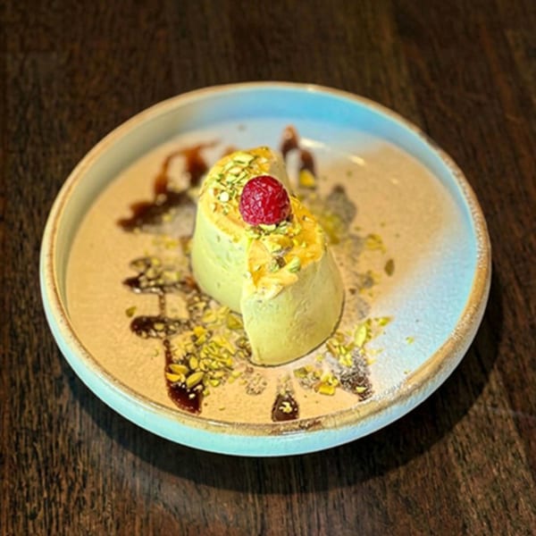 Pistachio dessert with a raspberry on top, drizzled with chocolate sauce on a light blue plate.