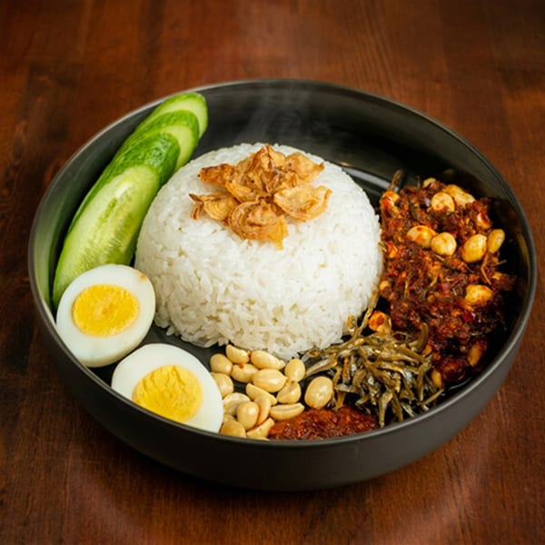 Nasi Lemak with rice, fried anchovies, peanuts, sambal, cucumber, and boiled eggs in a black bowl.