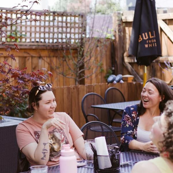 Three women laughing and talking at an outdoor patio table with a wooden fence background.
