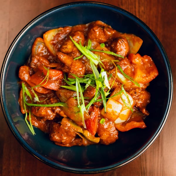 Close-up of chili chicken with red sauce, bell peppers, and green onions in a dark blue bowl.